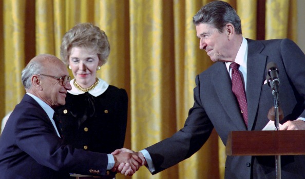 Milton Friedman shaking hands with Ronald Reagan while Nancy Reagan looks on. 