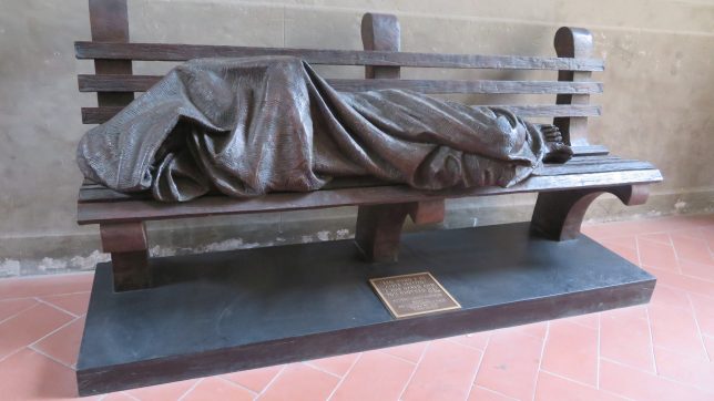 Bronze sculpture of a figure huddled in a blanket on a park bench. 